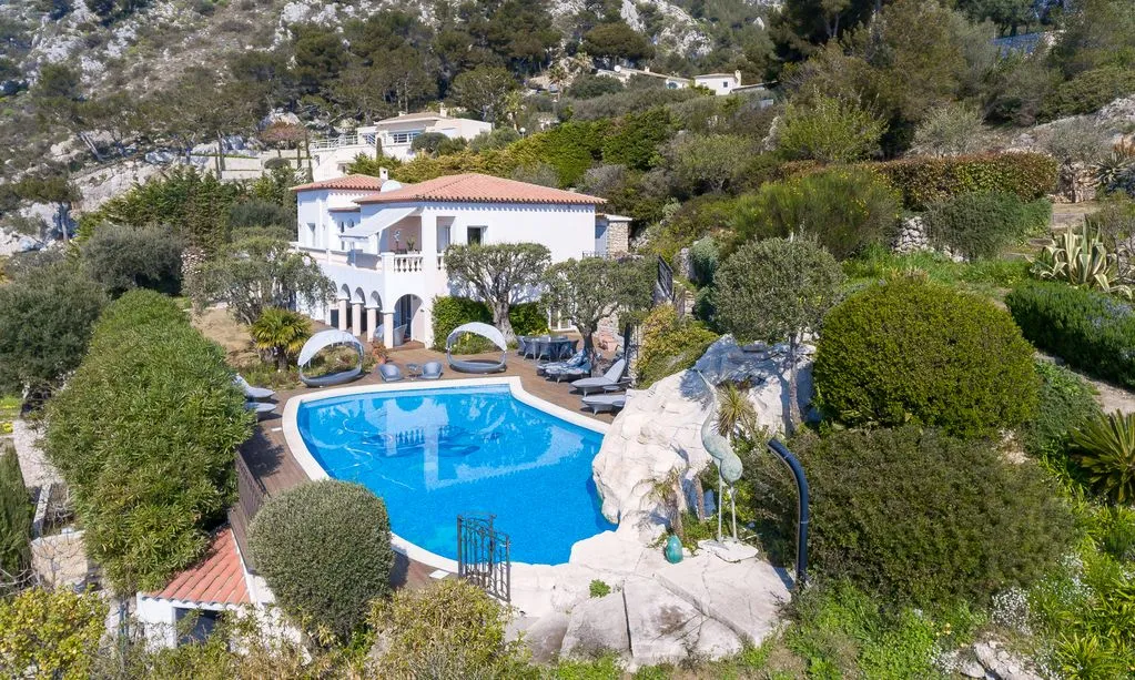 Modern two-story house with large glass windows, a pool, and surrounding trees; another building visible in the background.