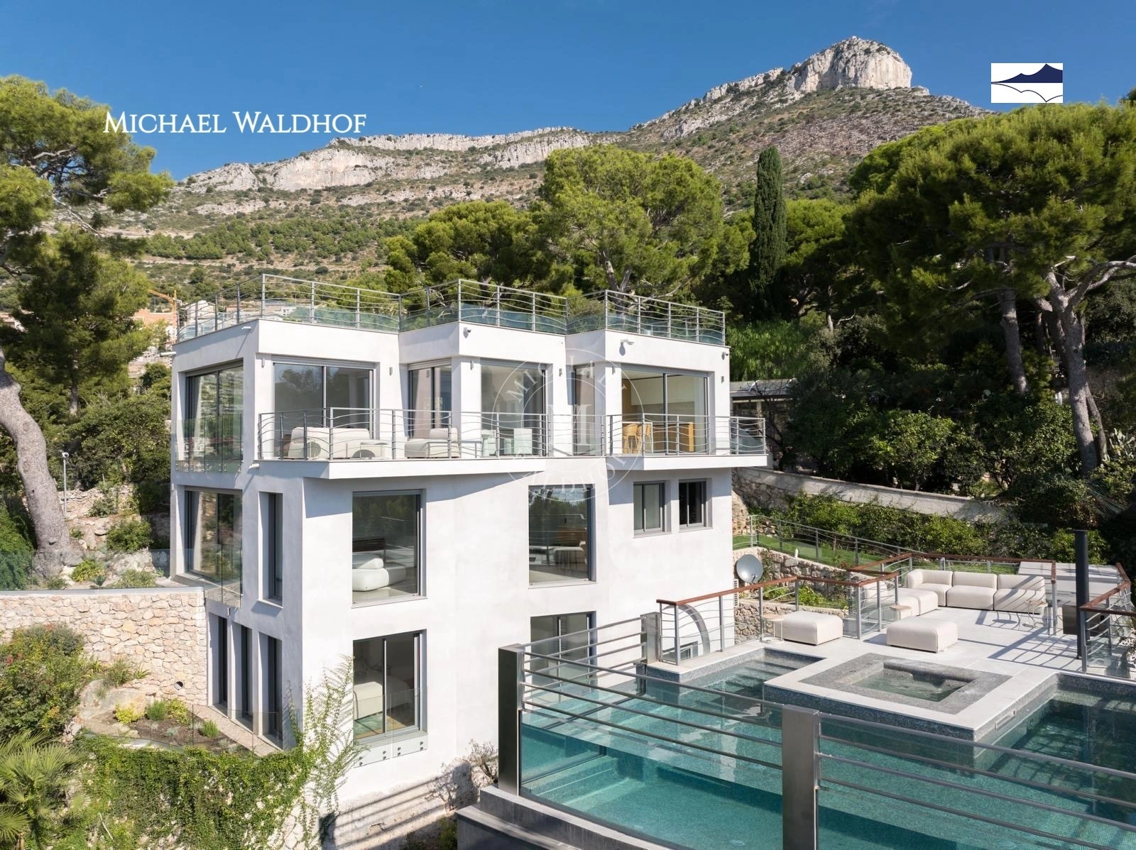 Modern white multi-story house with large windows, rooftop terraces, and a pool, set on a hillside with trees and mountains in the background.